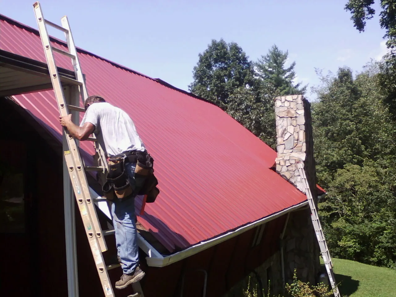 Roofer on a ladder beside a red metal roof and stone chimney — Soffit & Fascia Repair by Roofing Brothers in Tie Siding